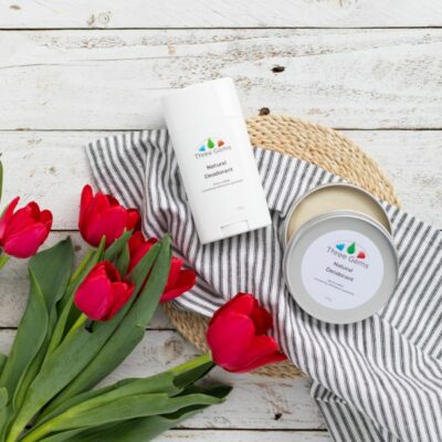 2 Varieties Of Three Gems Natural Deodorant Laying On Fabric With Red Tulips